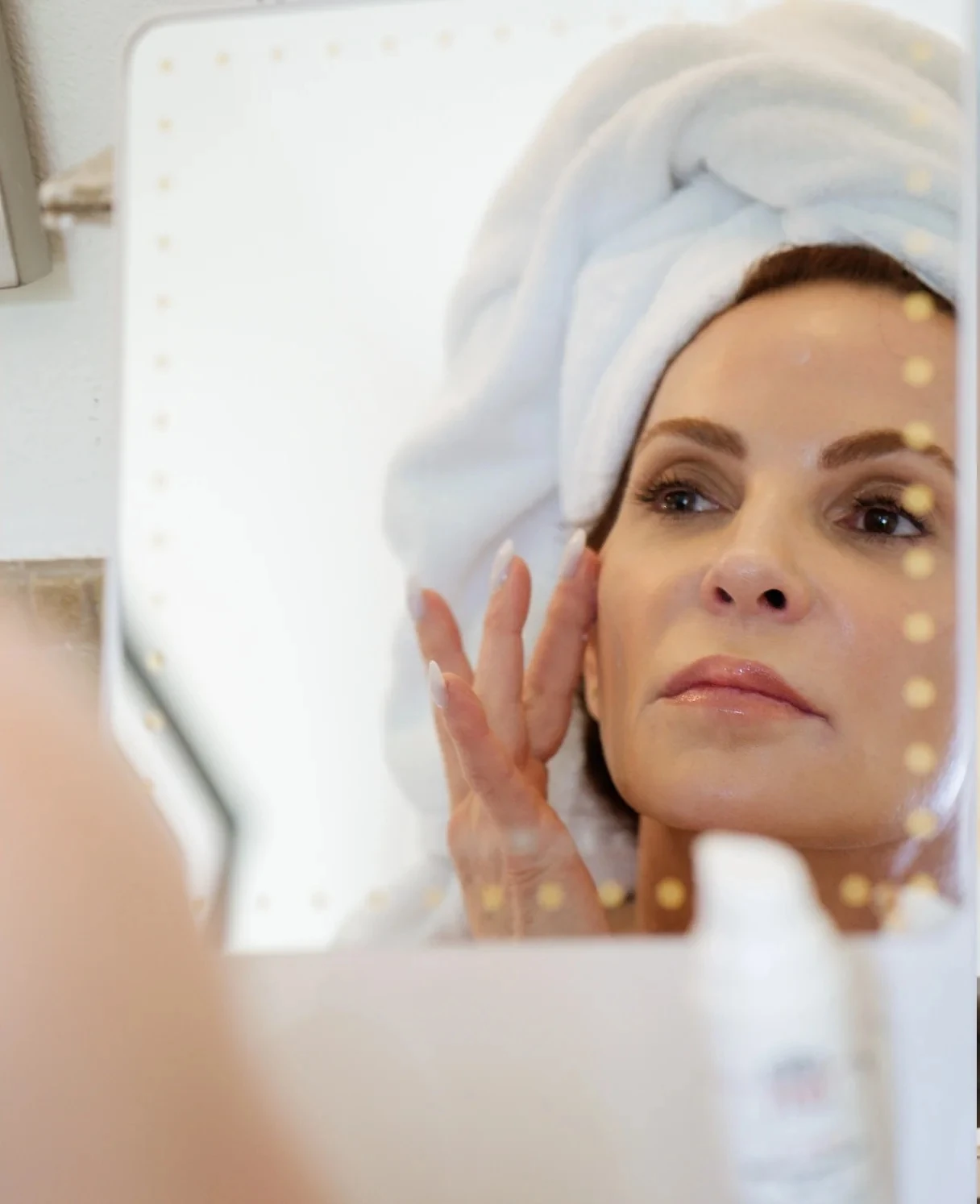 A woman with her hair wrapped in a towel, touching her cheek at a lit mirror